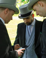 3 Models wear Black Tailcoat, Powder Blue Waistcoat & Grey Stripe Trousers Hire Package with Top Hat grouped together looking at one phone