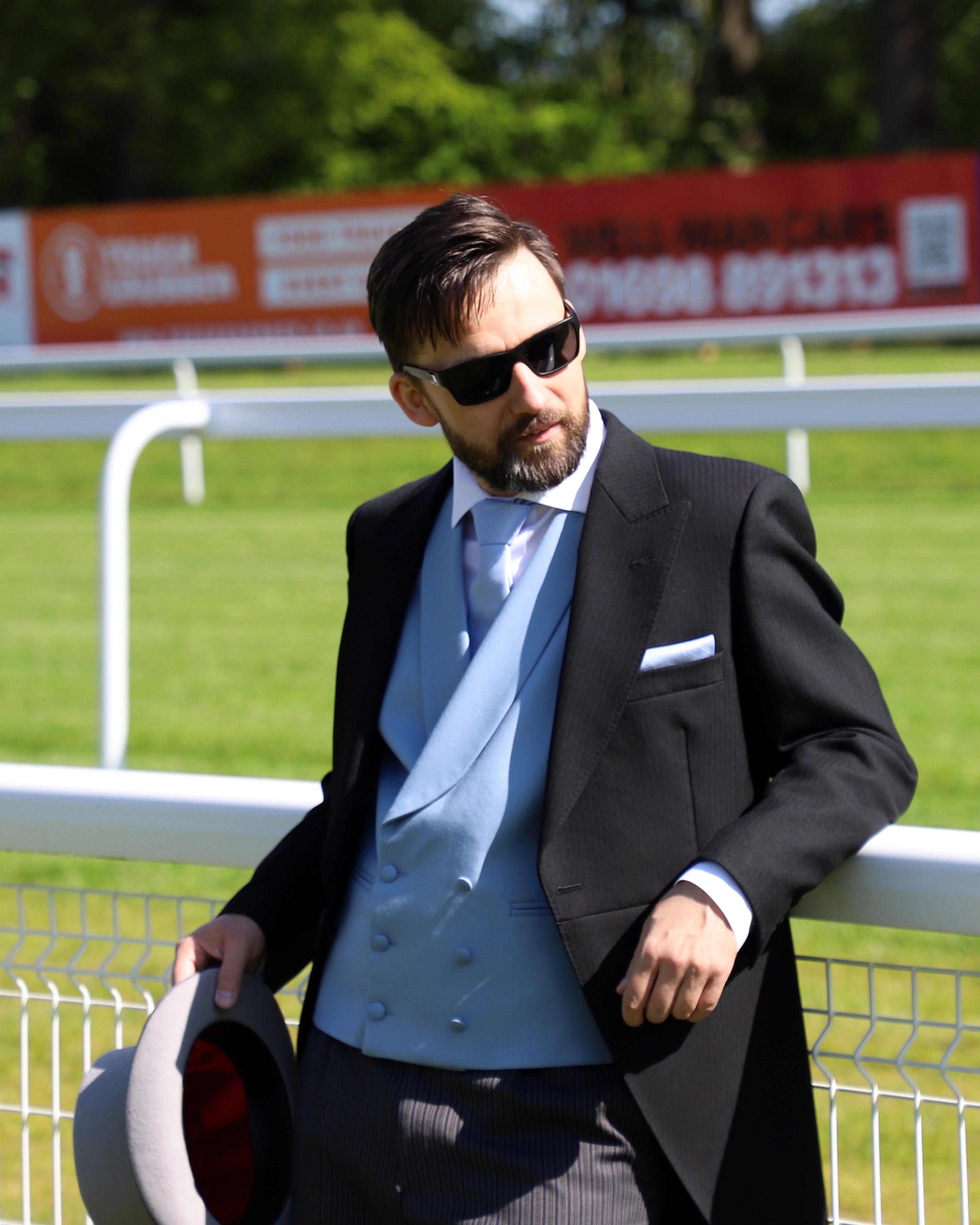 Model wears Black Tailcoat, Blue Waistcoat & Grey Stripe Trousers Hire Package with Top Hat leaning back on white rail