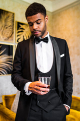 Black slim fit tuxedo with bow tie, model holding drink in relaxed pose.
