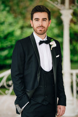 Groom wearing black slim fit tuxedo with buttonhole, outdoor setting.