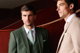 Two men in formal suits standing indoors. The man on the left wears a dark green three-piece suit with a white shirt and light green tie.