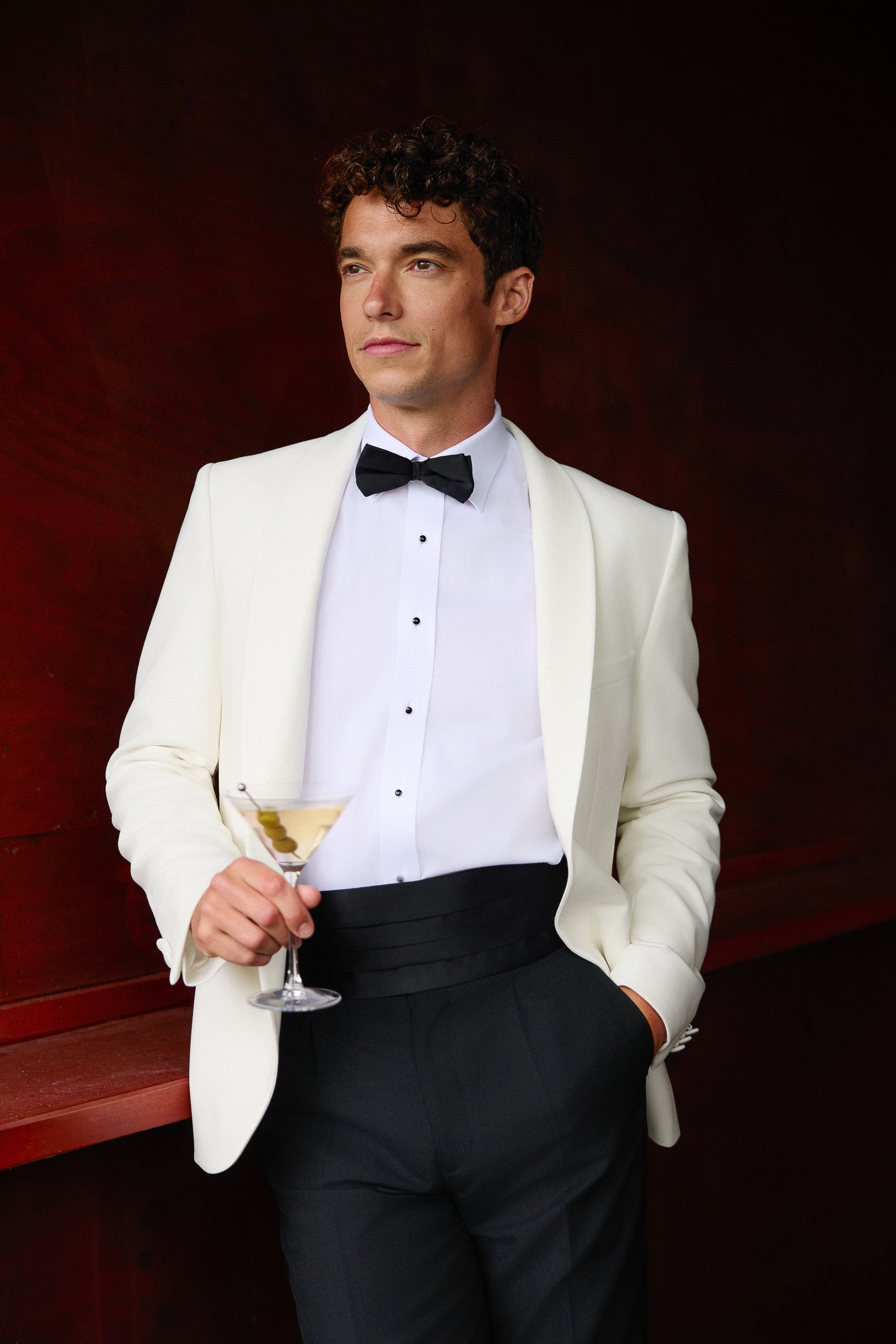 Man in a white tuxedo with a black bow tie holding a cocktail, sitting on a red bench against a dark background.