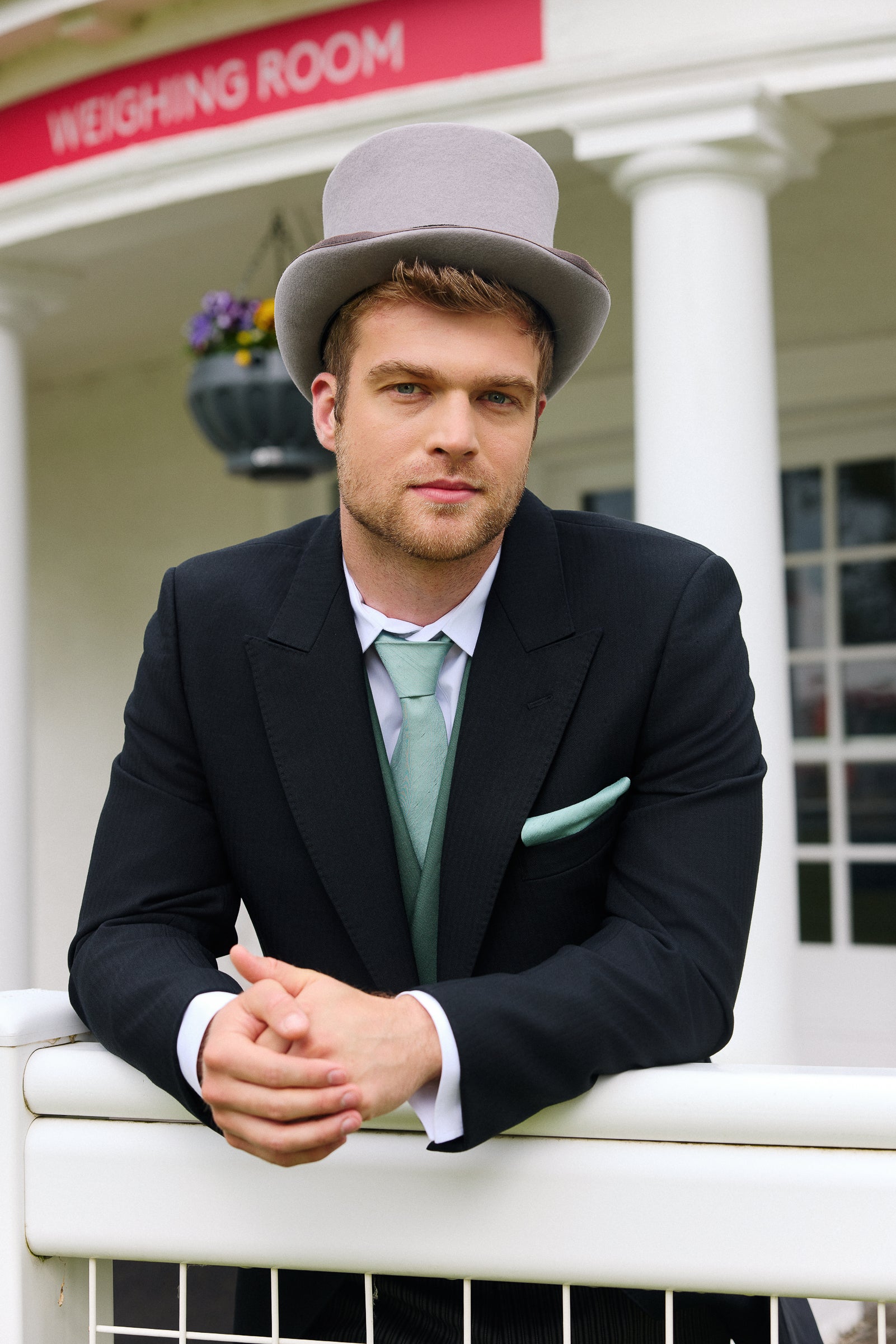 Man in Royal Ascot attire with a top hat leaning on a fence in front of a sign