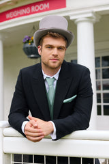 Man in Royal Ascot attire with a top hat leaning on a fence in front of a sign