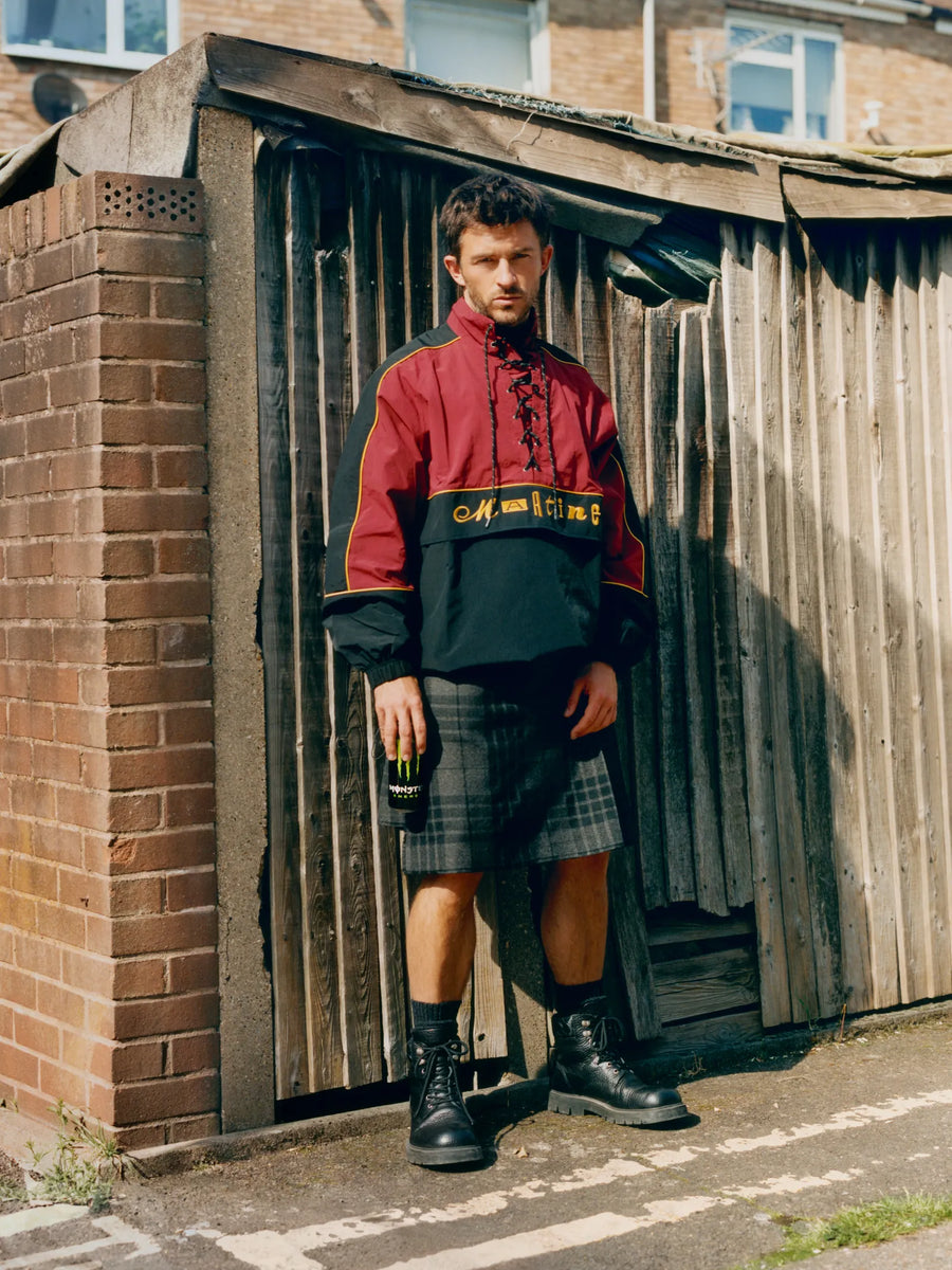 Johnathan Bailey standing wearing a red and black jacket with an ACS Grey Spirit Kilt standing in front of a wooden shed.