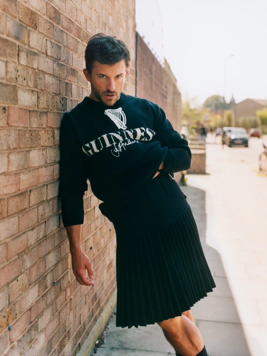 Johnathan Bailey wearing a black JW Anderson Guinness sweatshirt and ACS Grey Spirit Kilt leaning against a brick wall.
