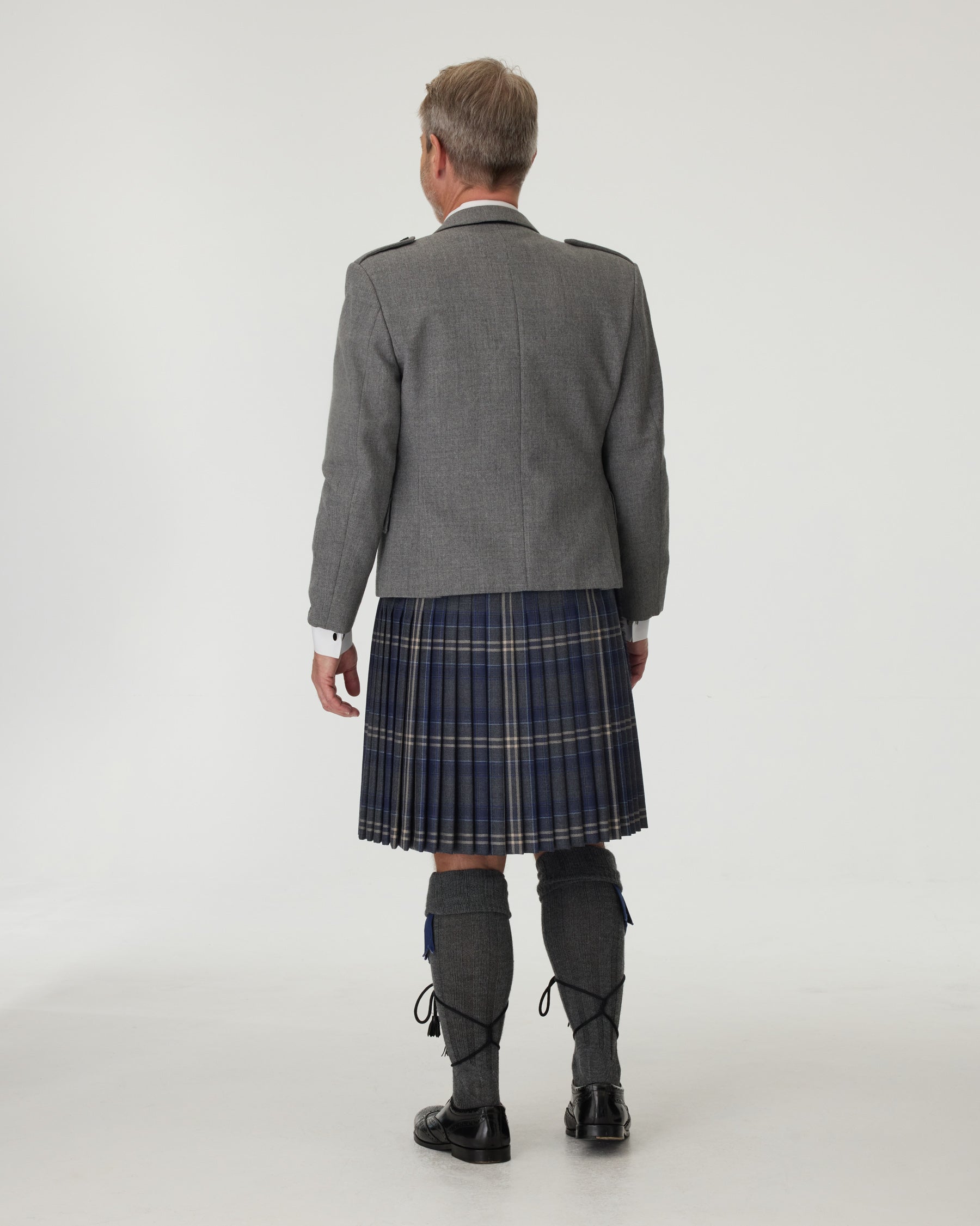 Man wearing a gray jacket and plaid kilt on a white background facing backwards